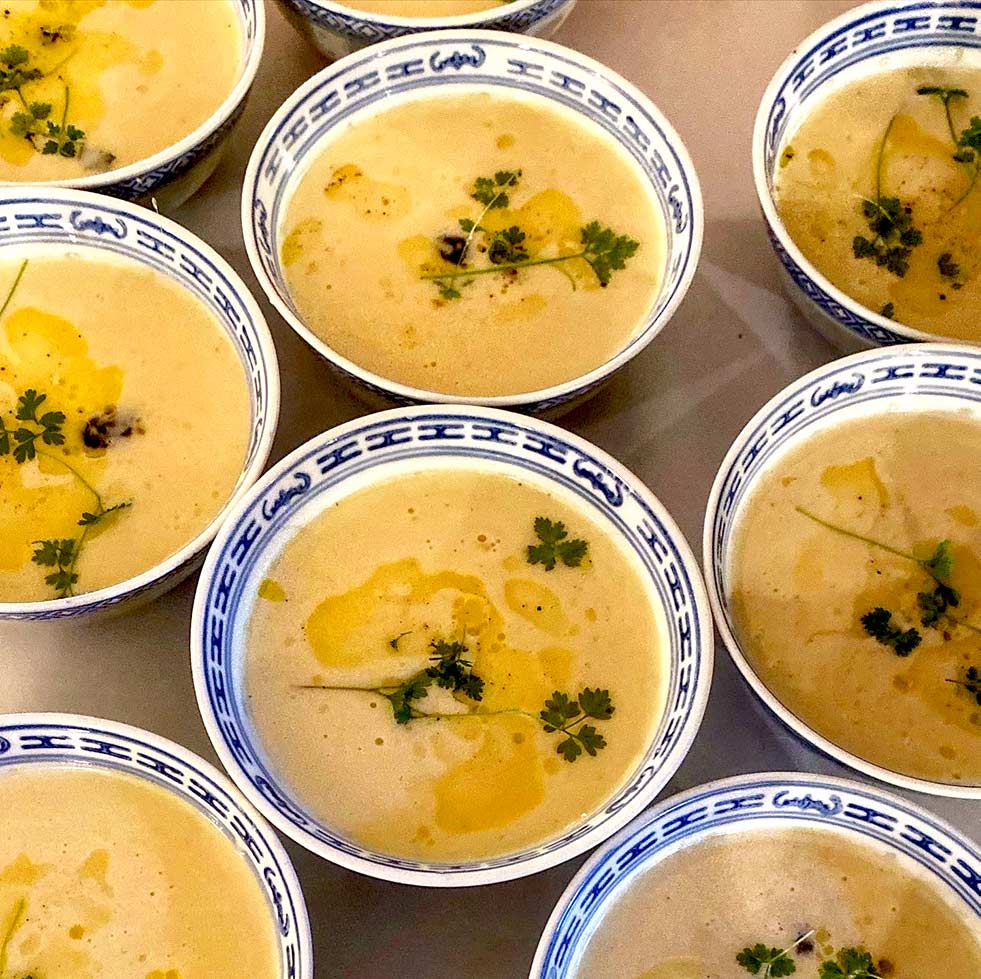 Several bowls of creamy yellow soup are garnished with fresh herbs and a drizzle of oil, served in blue and white patterned bowls, arranged closely together on a table.