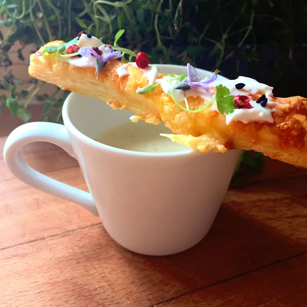A white cup filled with creamy soup is topped with a long, flaky pastry stick garnished with cream, microgreens, peppercorns, and edible flowers, set on a wooden surface with green plants in the background.