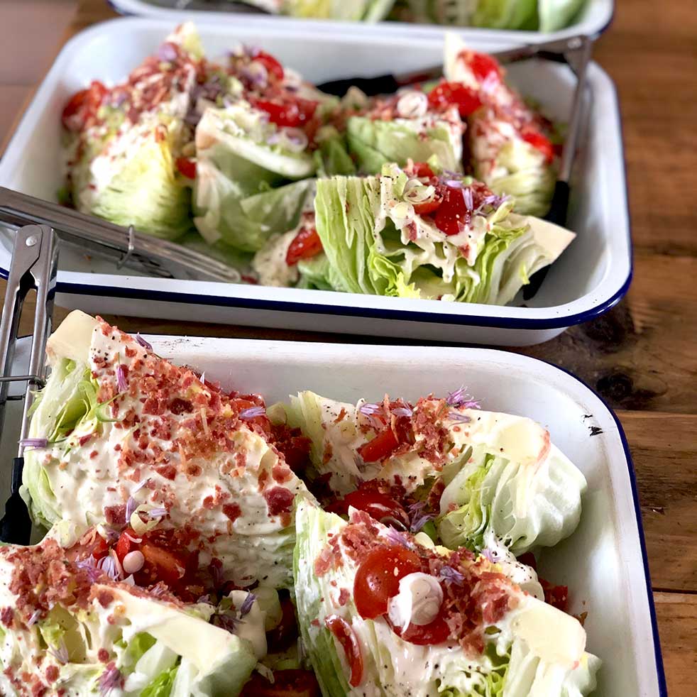 Trays of wedge salads topped with creamy dressing, diced tomatoes, bacon pieces, chopped red onion, and chives, served on white enamel trays with metal tongs on a wooden table.